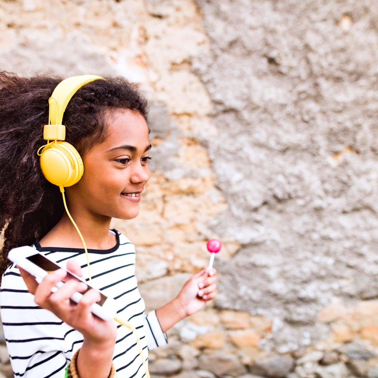 Beautiful african american girl with headphones, listening music Beautiful african american girl with curly hair outdoors, against stone wall, wearing headphones, listening music, eating lollipop.